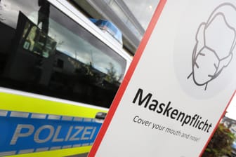 Ein Schild zur Maskenpflicht steht vor einem Polizeiauto (Symbolbild): In Wuppertal haben die Corona-Kontrollen der Stadt bisher Hunderttausende Euro Bußgeld eingebracht.