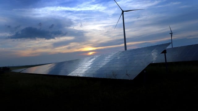 Der mit Wolken überzogene Abendhimmel spiegelt sich auf Solarzellen einer vor Windrädern stehenden Solarkraftanlage.