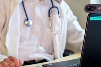 Ein Hausarzt sitzt während einer Videosprechstunde in seiner Praxis vor einem Laptop.