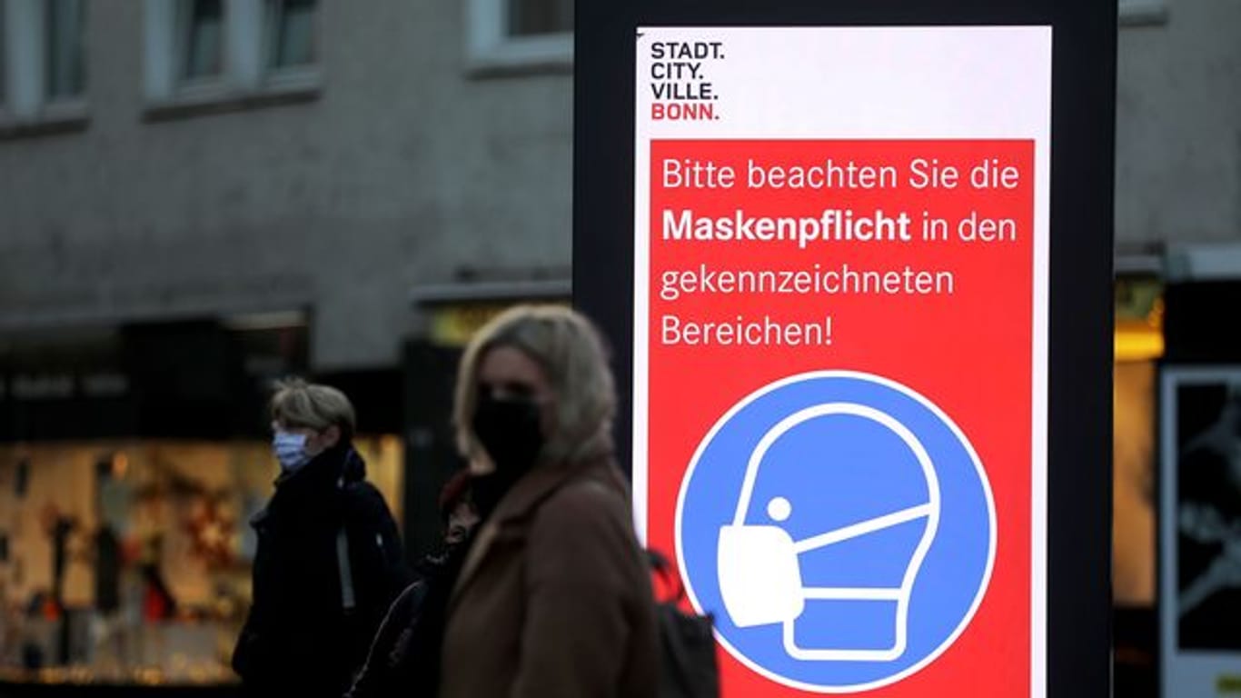 Frauen stehen neben einem Hinweisschild zur Maskenpflicht an einer Haltestelle in Bonn.