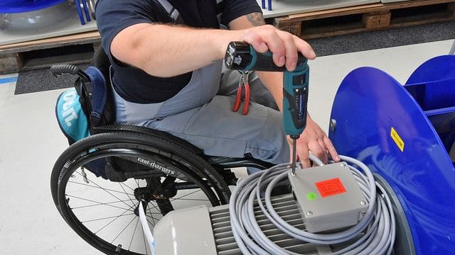 An einem Arbeitsplatz für Motorklemmen ist ein Mitarbeiter im Rollstuhl mit Montagearbeiten beschäftigt.
