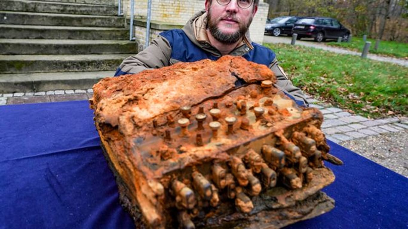 Florian Huber, Forschungstaucher, kniet vor dem archäologischen Landesamt Schleswig-Holstein bei der Übergabe der Enigma-Chiffriermaschine, die von ihm in der Ostsee gefunden wurde, neben der Maschine.