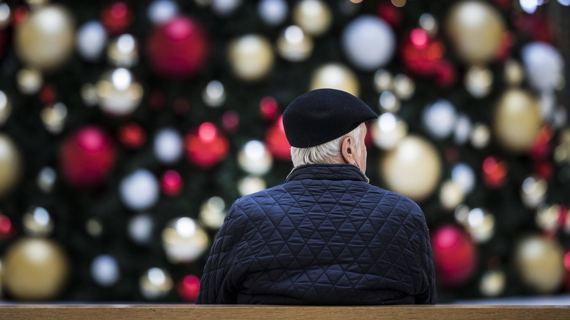 Ein Mann sitzt allein auf einer Bank vor einem geschmückten Weihnachtsbaum: Senioren haben Angst Weihnachten allein zu verbringen.