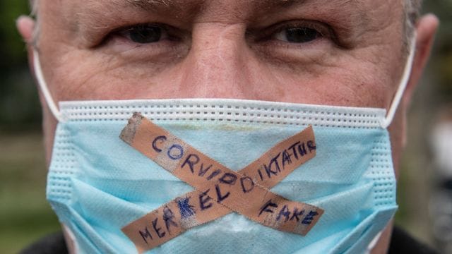 Ein Teilnehmer einer Kundgebung gegen die Corona-Maßnahmen der Regierung trägt einen Mundschutz mit der Aufschrift "Merkel-Diktatur - Covid-Fake". Ein Teilnehmer einer Kundgebung gegen die Corona-Maßnahmen der Regierung trägt einen Mundschutz mit der Aufschrift "Merkel-Diktatur - Covid-Fake".