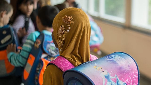 Das Symbolfoto zeigt ein Mädchen mit Kopftuch vor einem Klassenzimmer.