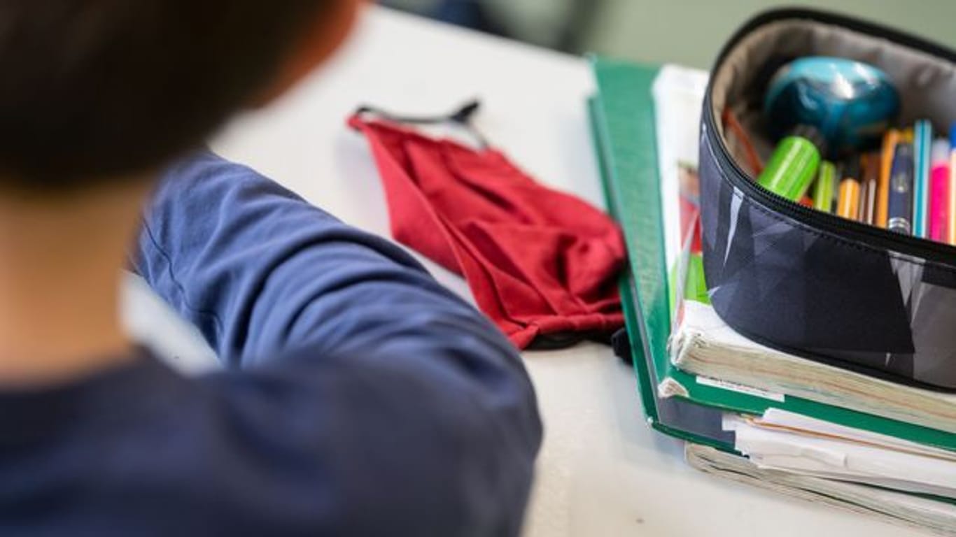 Eine Mund-Nasen-Bedeckung liegt während einer Unterrichtsstunde neben einem Mäppchen und Schulbüchern auf einem Schultisch.