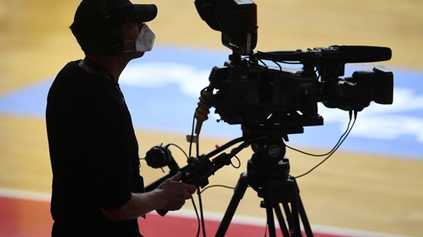 Ein Kameramann steht mit Mundschutz bei einem Basketballspiel an seiner TV-Kamera.