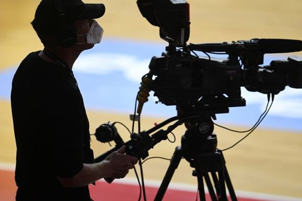 Ein Kameramann steht mit Mundschutz bei einem Basketballspiel an seiner TV-Kamera.