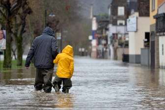 Zwei Menschen in Wathosen schlendern durch die von der Mosel überflutete Uferstraße der Gemeinde Reil: Tauwasser und Regen sorgen für steigende Pegelstände.
