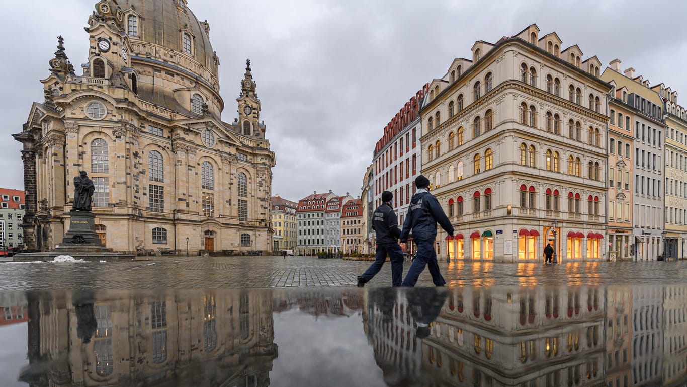 Dresden: Die Frauenkirche und die Häuser auf dem Neumarkt spiegeln sich am Nachmittag in einer nassen Steinbank während zwei Mitarbeiter der Polizeibehörde (Ordnungsamt) entlang laufen.