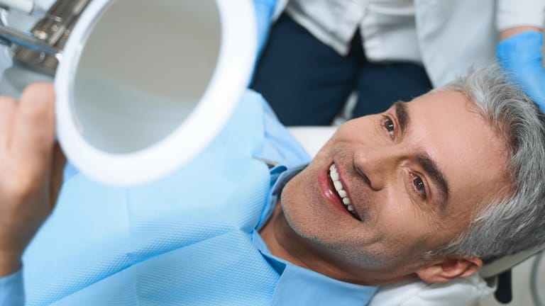 Ein Patient auf einer Behandlungsliege betrachtet das Resultat seiner Zahnbehandlung. Beim Zahnersatz müssen Qualität und Optik stimmen.