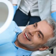 Ein Patient auf einer Behandlungsliege betrachtet das Resultat seiner Zahnbehandlung. Beim Zahnersatz müssen Qualität und Optik stimmen.
