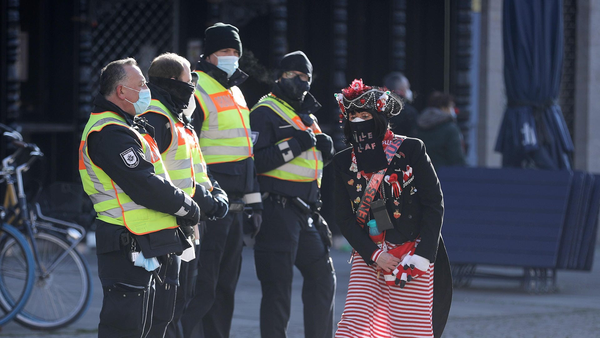 Eine Karnevalistin tanzt an Weiberfastnacht auf dem Alter Markt neben Mitarbeitern des Ordnungsamtes. Wegen Corona fällt der Straßenkarneval in Nordrhein-Westfalen aus, trotzdem wurde viele Verstöße geahndet.