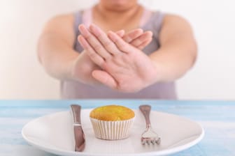 Fastenzeit: Auf einem Teller liegt ein Muffin. Eine Person sitzt davor und hält abweisend die Hände zwischen sich und den Teller.