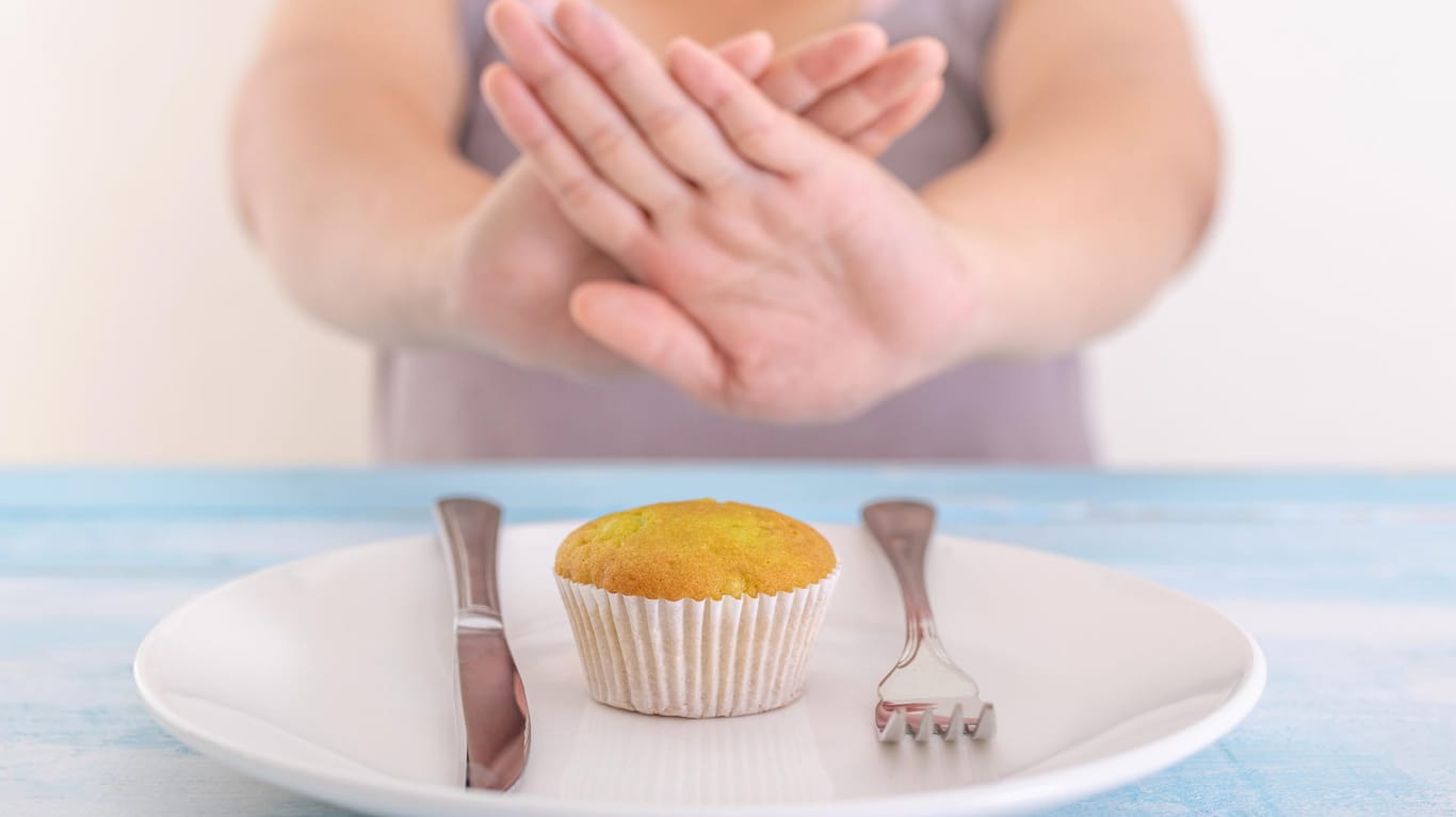 Fastenzeit: Auf einem Teller liegt ein Muffin. Eine Person sitzt davor und hält abweisend die Hände zwischen sich und den Teller.