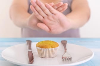 Fastenzeit: Auf einem Teller liegt ein Muffin. Eine Person sitzt davor und hält abweisend die Hände zwischen sich und den Teller.