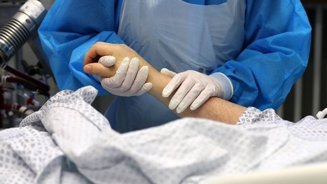 Im Infektionszimmer hält die Mitarbeiterin einer Intensivstation die Hand eines Covid-19-Patienten: Laut dem Chef-Pathologen der Uniklinik Augsburg sind die meisten Corona-Toten an und nicht mit dem Virus gestorben.