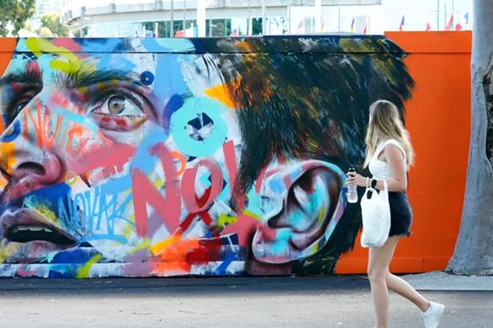 Eine Frau geht an einem Wandbild von Novak Djokovic vorbei, das auf eine Wand im Melbourne Park gemalt wurde.