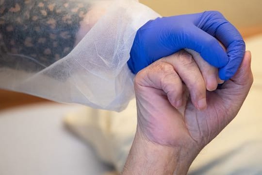 Eine Altenpflegerin in Schutzkleidung hält die Hand eines Bewohners.