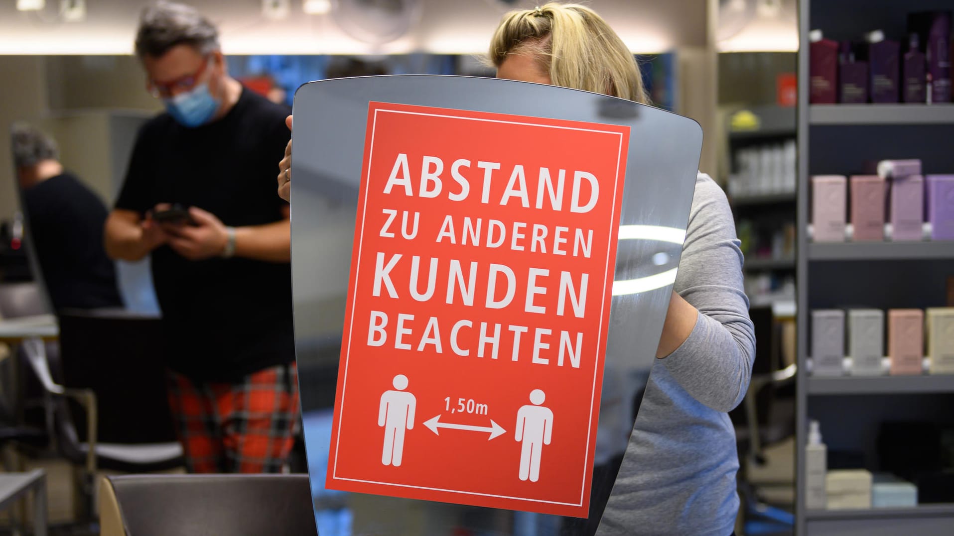 Eine Friseur-Mitarbeiterin stellt einen Spiegel mit der Aufschrift "Abstand zu anderen Kunden beachten" auf einen Tisch.