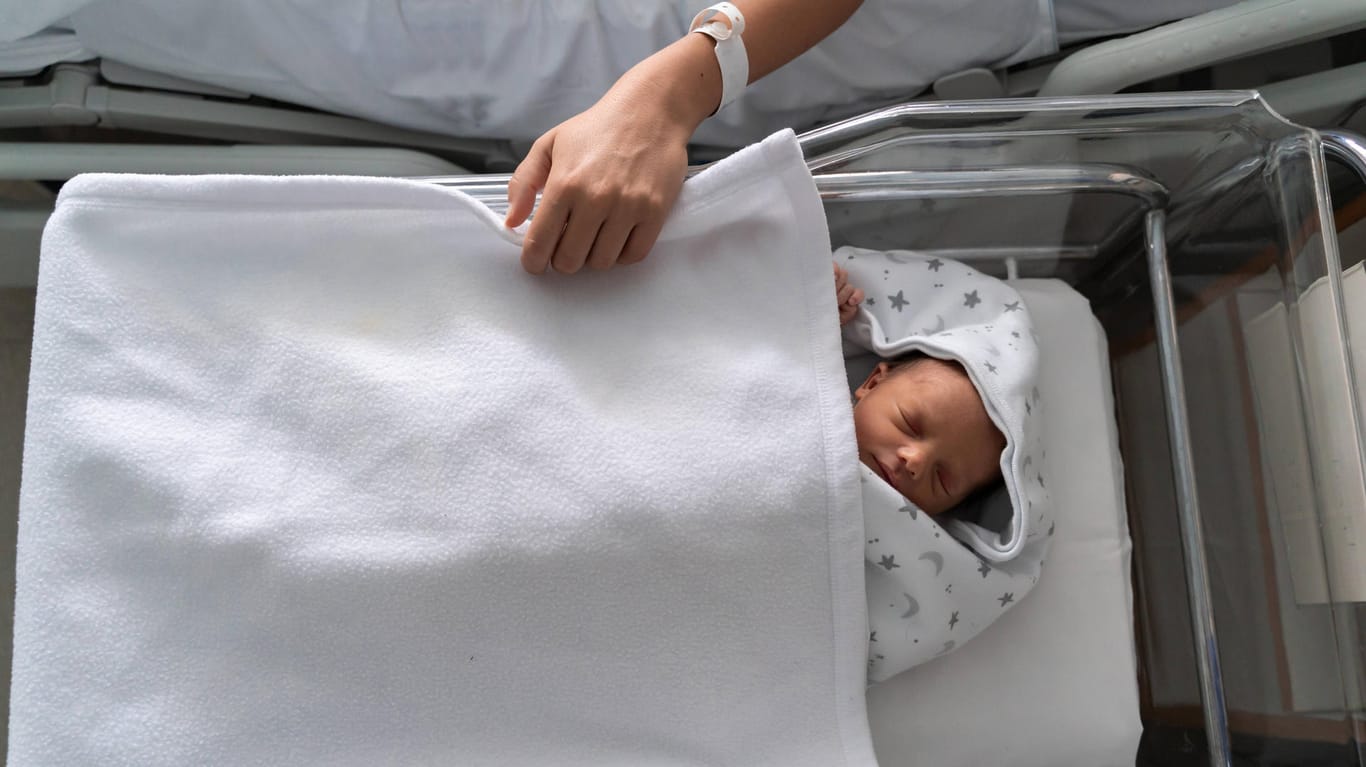 Hand einer Mutter auf dem Bett eines Neugeborenen (Symbolbild): In Großbritannien infizierte sich eine Frau in der Schwangerschaft mit Corona, ihr Kind sah sie erst jetzt.