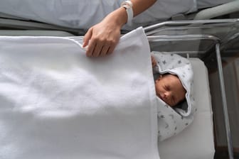 Hand einer Mutter auf dem Bett eines Neugeborenen (Symbolbild): In Großbritannien infizierte sich eine Frau in der Schwangerschaft mit Corona, ihr Kind sah sie erst jetzt.