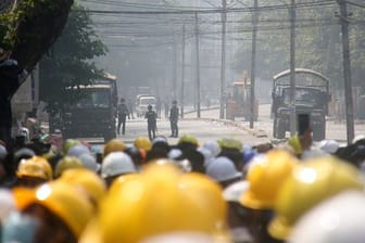 Demonstranten in Mandalay tragen gelbe Schutzhelme und beobachten auf einer blockierten Straße die Polizei.