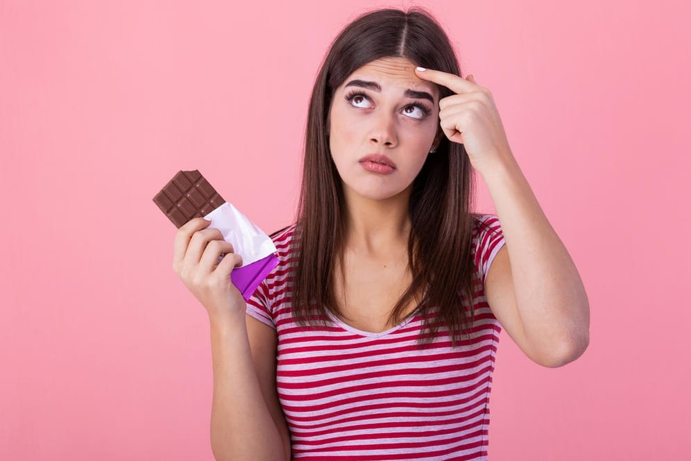 Eine junge Frau mit einer Tafel Schokolade blickt skeptisch empor: Zucker steht im Verruf, der Haut zu schaden.