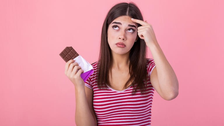Eine junge Frau mit einer Tafel Schokolade blickt skeptisch empor: Zucker steht im Verruf, der Haut zu schaden.