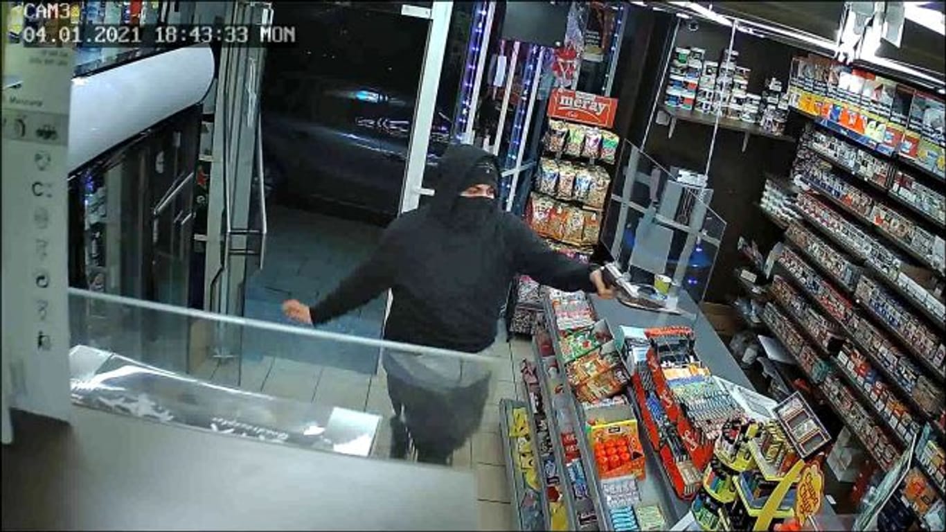 Ein Mann richtet eine Pistole in einem Kiosk auf eine Person: Die Polizei fahndet mit diesem Foto nach dem Kiosk-Räuber.