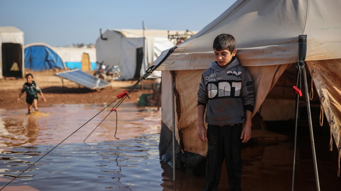 Syrien, Kafr Aruq: Ein syrischer Junge steht in einem Flüchtlingslager, das vom Regen überflutet wurde, inmitten des schlammigen Wassers vor seinem Zelt.