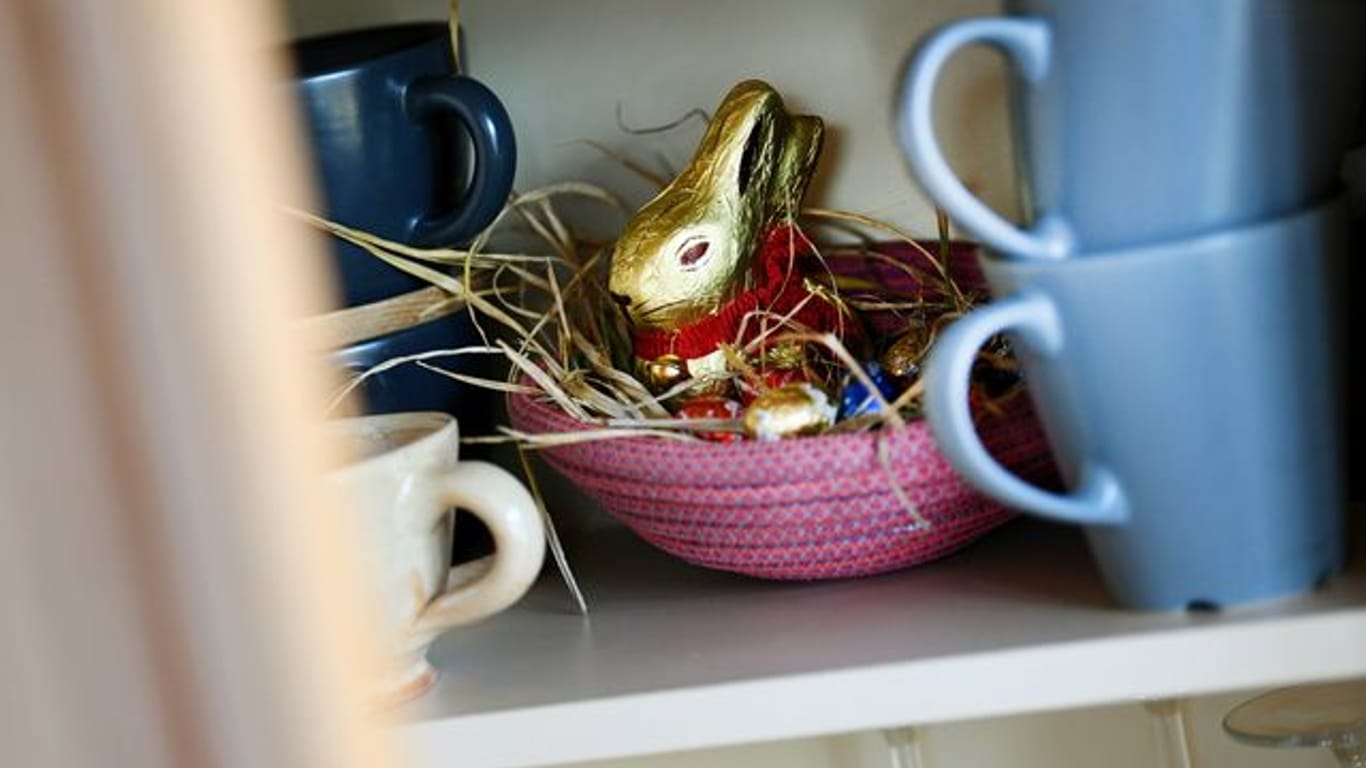 Ein Osternest mit Schokohase und Schokoladeneiern ist in einer Wohnung zwischen Geschirr im Schrank versteckt.