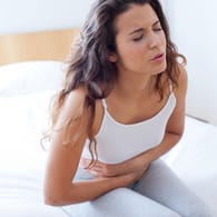 Eine Frau mit Bauchkrämpfen sitzt auf dem Bett: Wiederkehrende Schmerzen im Oberbauch deuten auf einen Reizmagen hin, werden aber oft als Gastritis fehlgedeutet,