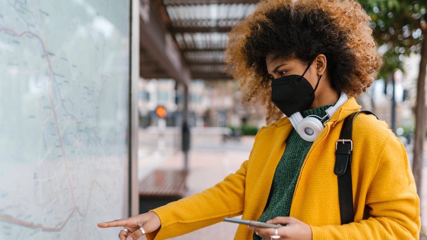 Eine Frau mit Maske und Smartphone (Symbolbild): Die Kölner App "Darf ich das?" will einen bundesweiten Überblick zu den aktuellen Corona-Regeln bieten.