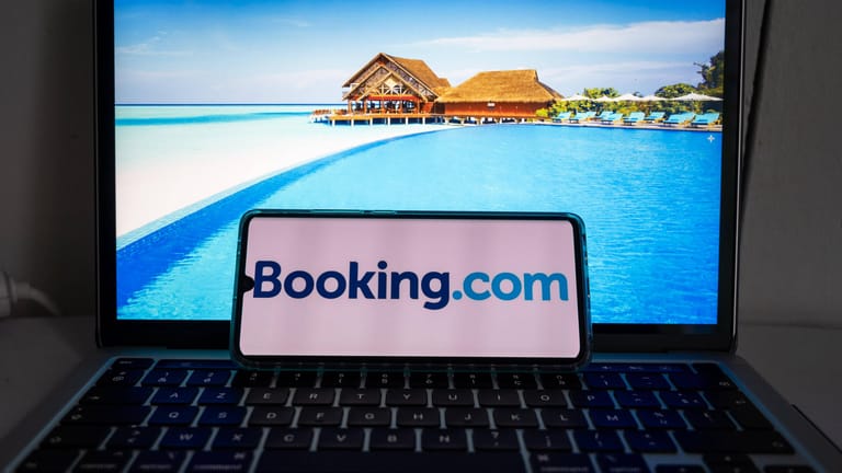 Ein Smartphone mit dem Logo von "Booking.com" liegt auf einem Laptop mit Urlaubsbildern: Das Urlaubsportal hat eine Datenpanne zu spät an die zuständigen Behörden gemeldet.