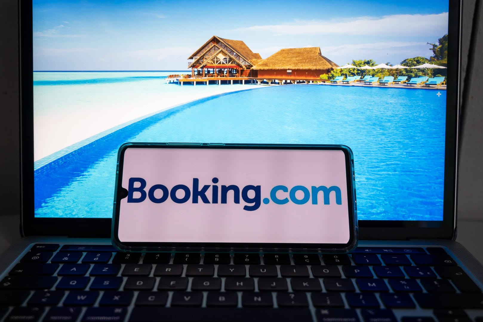 Ein Smartphone mit dem Logo von "Booking.com" liegt auf einem Laptop mit Urlaubsbildern: Das Urlaubsportal hat eine Datenpanne zu spät an die zuständigen Behörden gemeldet.