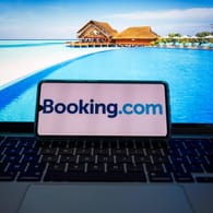 Ein Smartphone mit dem Logo von "Booking.com" liegt auf einem Laptop mit Urlaubsbildern: Das Urlaubsportal hat eine Datenpanne zu spät an die zuständigen Behörden gemeldet.