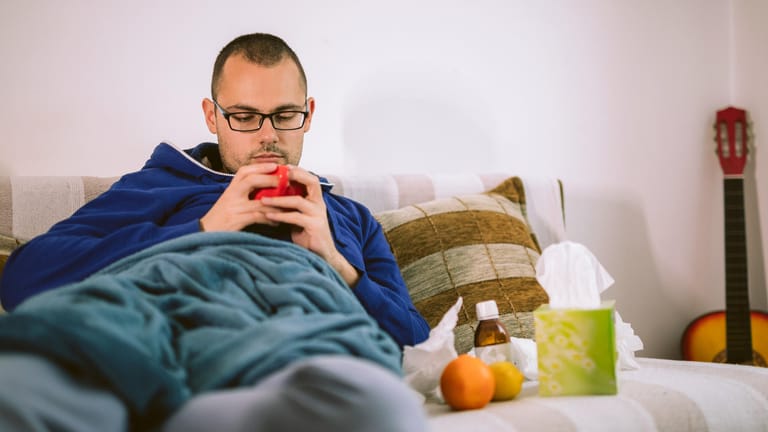 Ein Mann liegt krank im Bett: Typische Symptome des Pfeifferschen Drüsenfiebers sind geschwollene Lymphknoten, Halsschmerzen und Fieber.