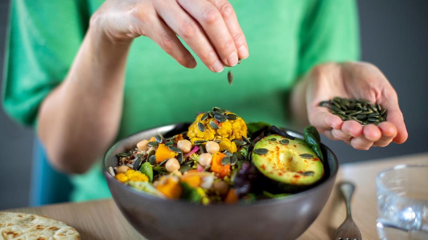 Eine Frau streut Kürbiskerne in eine vegane Bowl. Wer sich ohne tierische Produkte ernährt, sollte ein paar wichtige Grundregeln beachten.