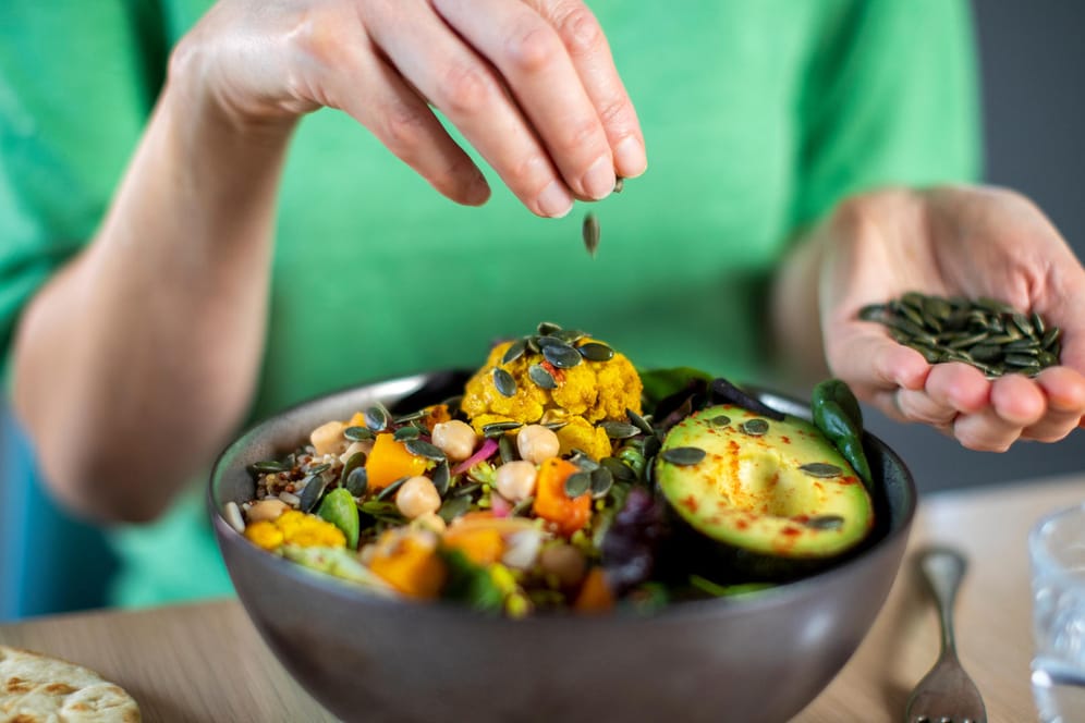 Eine Frau streut Kürbiskerne in eine vegane Bowl. Wer sich ohne tierische Produkte ernährt, sollte ein paar wichtige Grundregeln beachten.