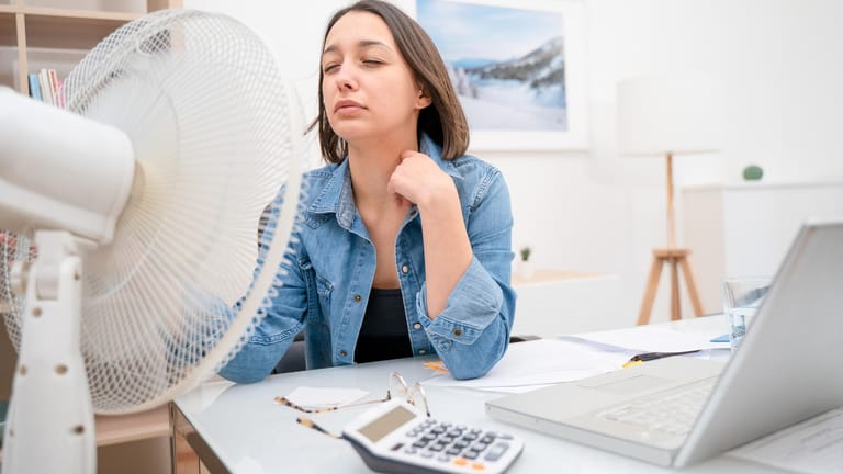 Frau sitzt vor einem Ventilator: Einige Tipps und Tricks können helfen, bestmöglich durch die Hitze zu kommen.