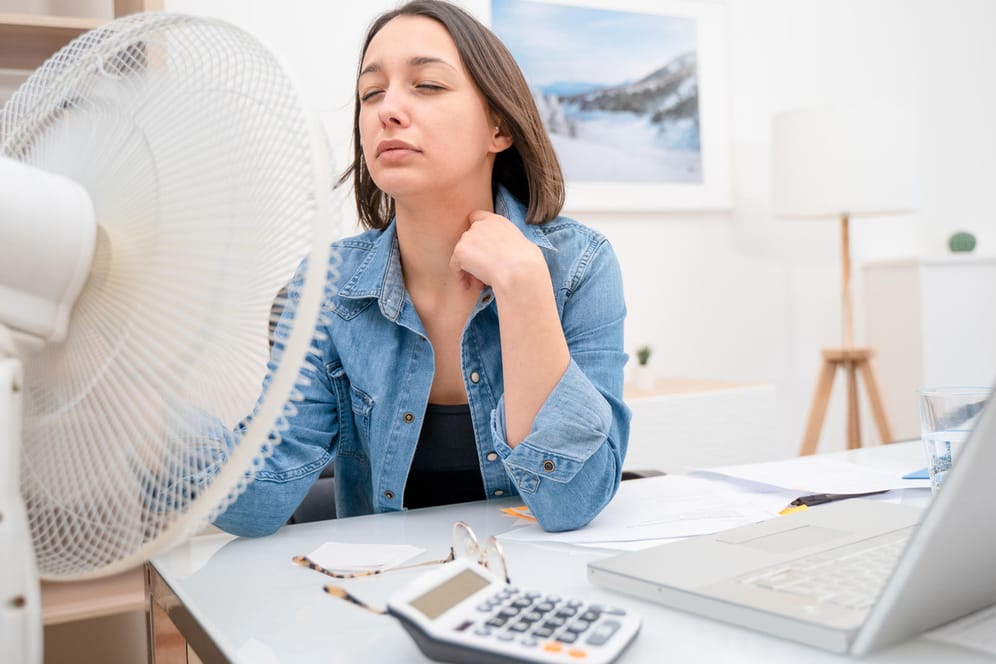 Frau sitzt vor einem Ventilator: Einige Tipps und Tricks können helfen, bestmöglich durch die Hitze zu kommen.