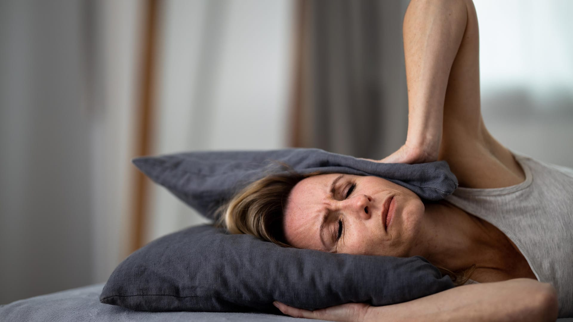 Eine Frau mit gequältem Gesichtsausdruck bedeckt die Ohren mit einem Kissen. Die störenden Ohrgeräusche bei Tinnitus können sehr belastend sein. Häufig entstehen sie durch Dauerstress.