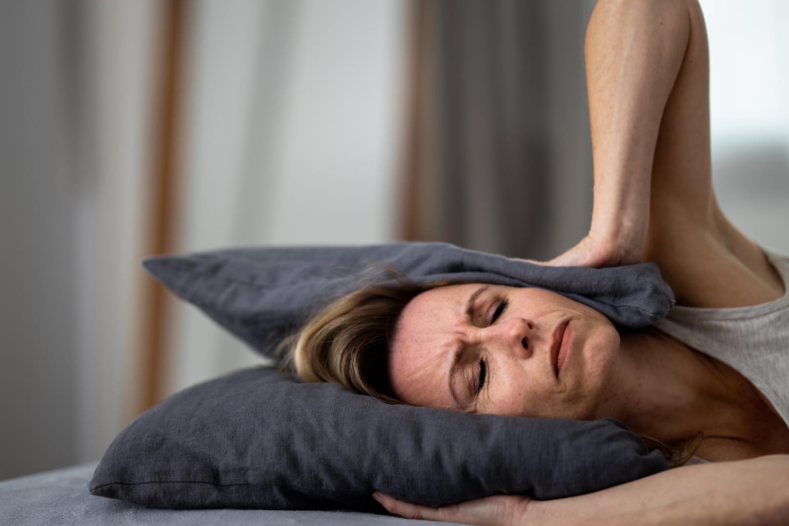 Eine Frau mit gequältem Gesichtsausdruck bedeckt die Ohren mit einem Kissen. Die störenden Ohrgeräusche bei Tinnitus können sehr belastend sein. Häufig entstehen sie durch Dauerstress.