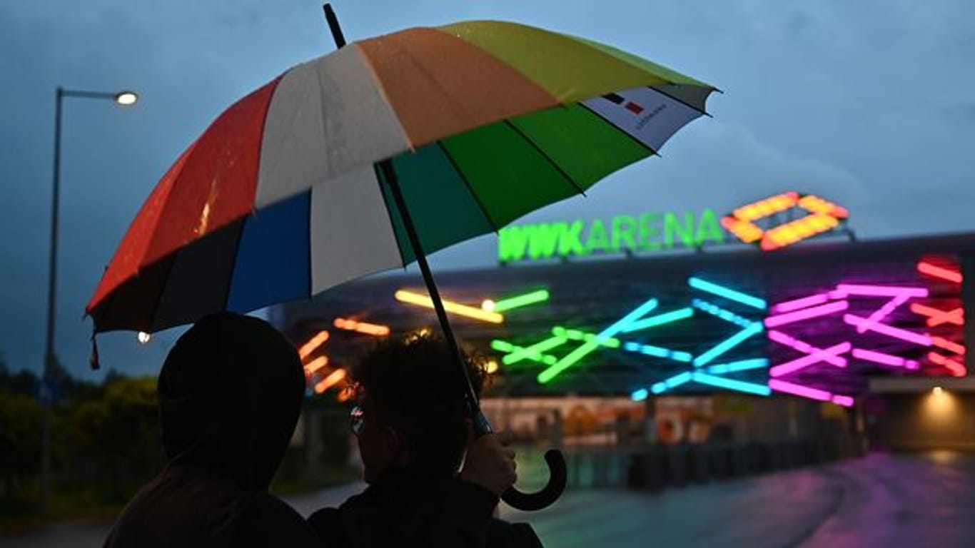 FußZwei junge Männer mit einem Regenbogen-Schirm stehen vor WWK-Arena in Augsburg, die während des Spiels in Regenbogenfarben beleuchtet ist.