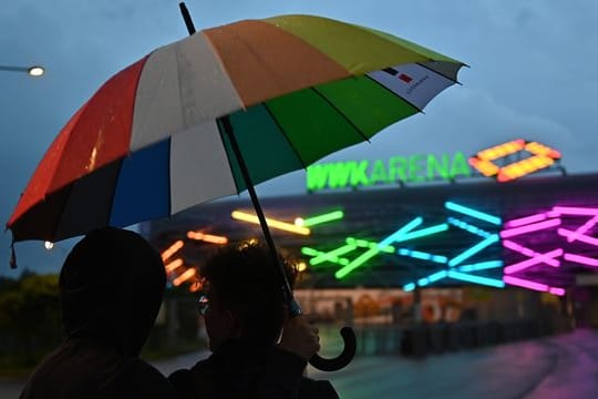 FußZwei junge Männer mit einem Regenbogen-Schirm stehen vor WWK-Arena in Augsburg, die während des Spiels in Regenbogenfarben beleuchtet ist.