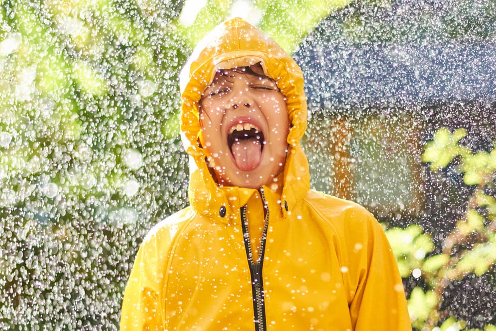 Ein fröhlicher Junge steht im Regen während im Hintergrund die Sonne scheint.