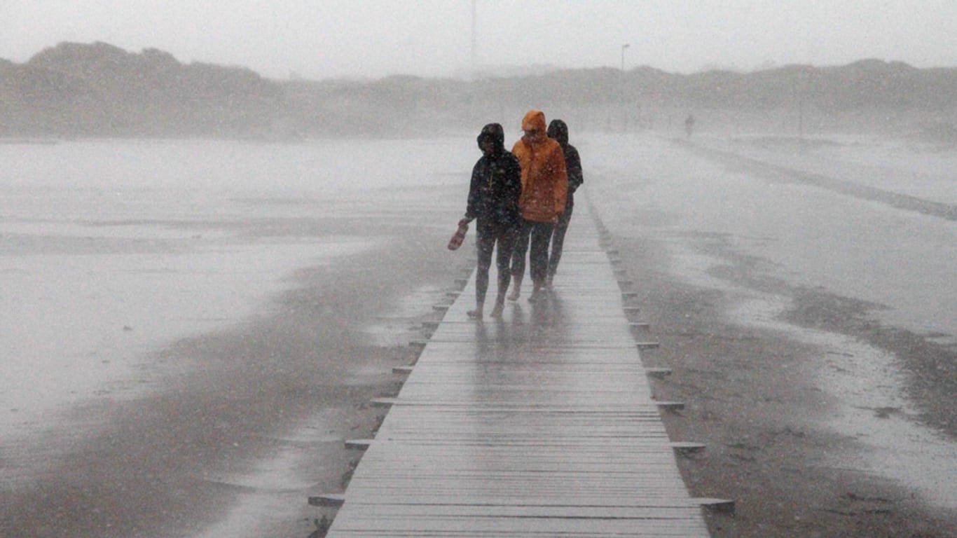 Von Sommer keine Spur: Spaziergänger gehen bei Sturm und heftigem Regen über einen Holzweg am Strand von St. Peter-Ording