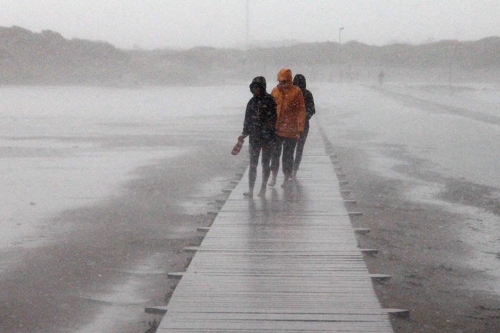 Von Sommer keine Spur: Spaziergänger gehen bei Sturm und heftigem Regen über einen Holzweg am Strand von St. Peter-Ording