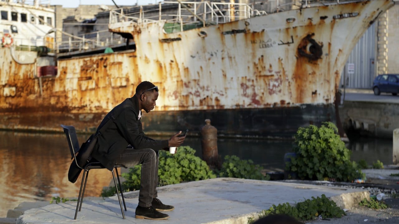 Ein Migrant mit seinem Mobiltelefon vor einem verrosteten Schiff im Hafen von Valletta.